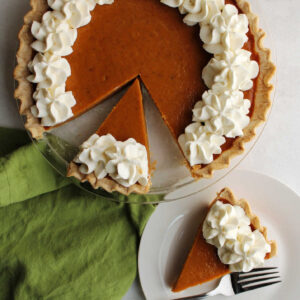 Piece of pumpkin pie topped with swirls of whipped cream next to remaining pie, ready to eat.