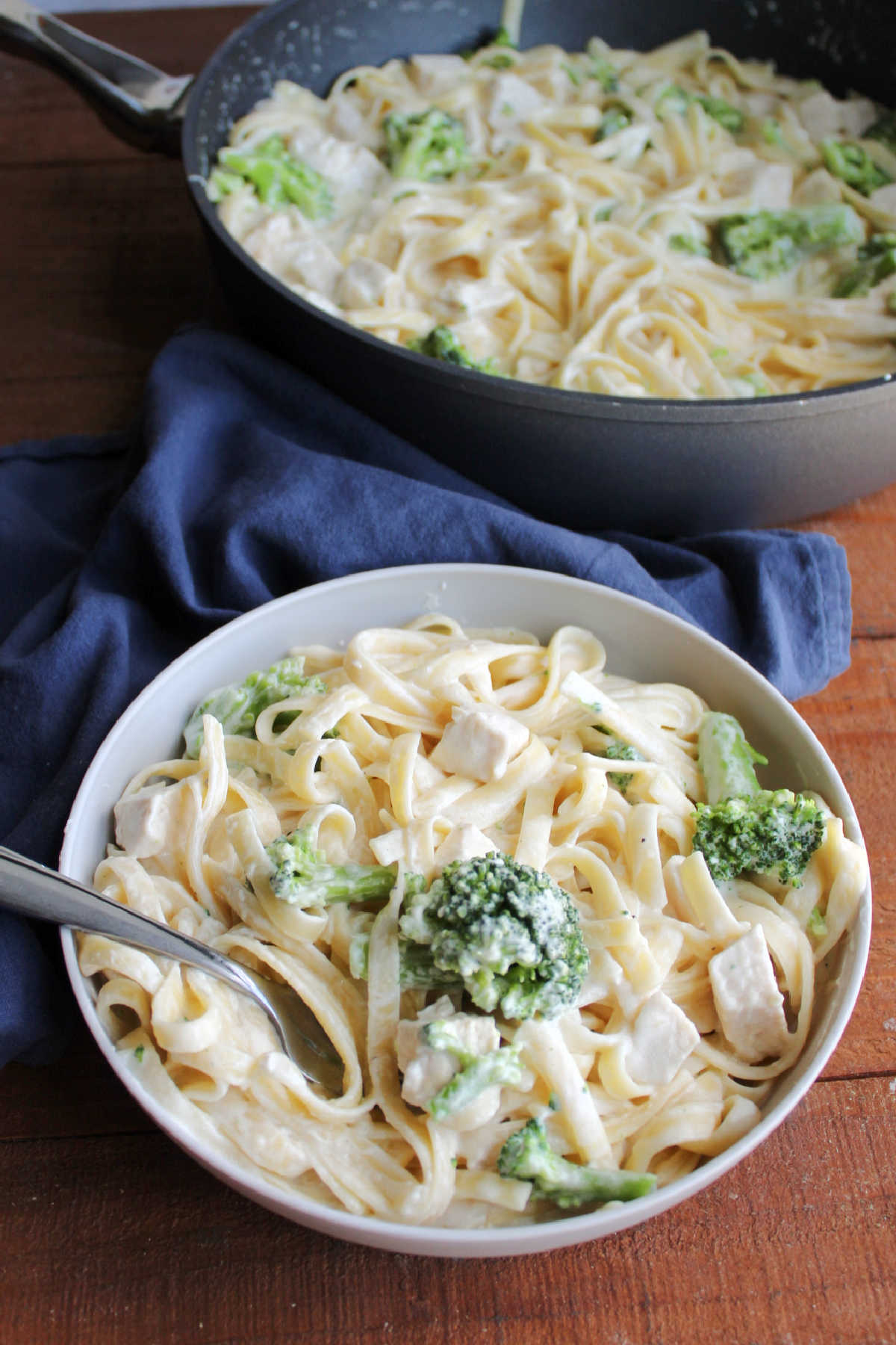 Serving of chicken, broccoli, and fettuccine coated in homemade alfredo sauce with pan and remaining pasta in the background. 