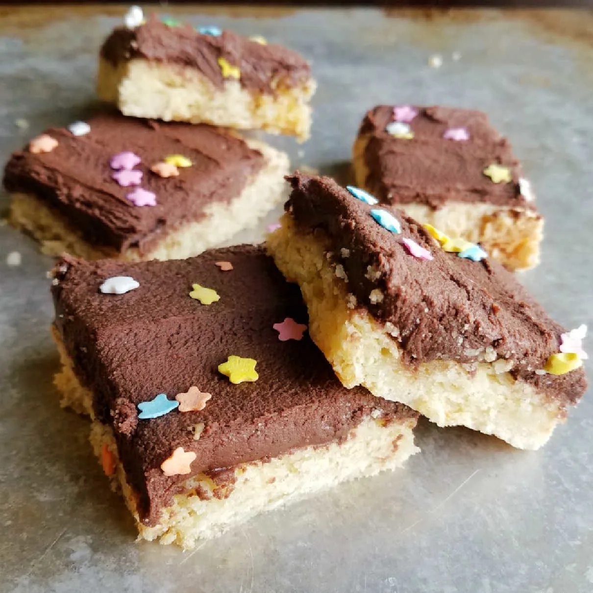 square pieces of sugar cookie bars with chocolate frosting and star sprinkles on top piled on baking sheet.