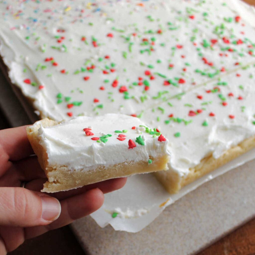 hand holding soft sugar cookie bar topped with fluffy white frosting and sprinkles.