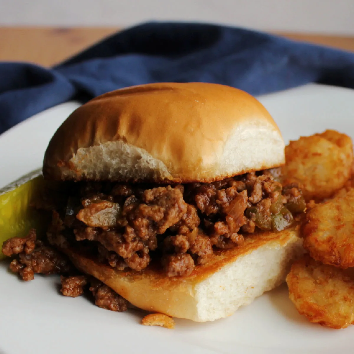 toasted bun filled with crumbly sloppy joe mixture with tater tots and a pickle on the side.