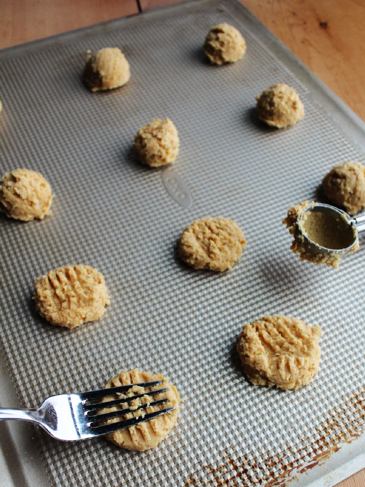 Tray of peanut butter cookies with cookie scoop nearby and a fork making the marks on the top of a ball of peanut butter cookie dough.