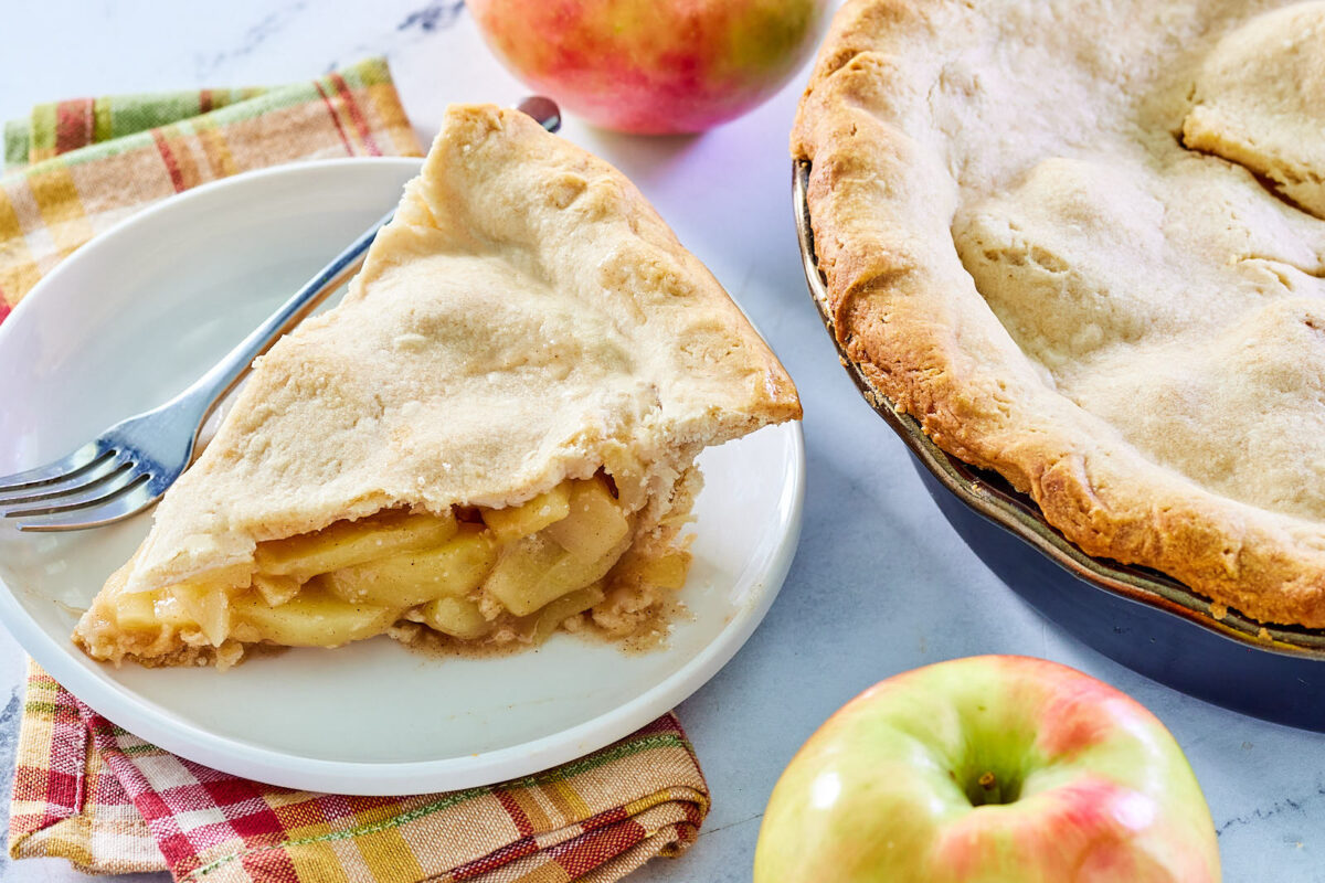 Piece of grandma's deep dish apple pie next to a couple of fresh apples and the rest of the pie.