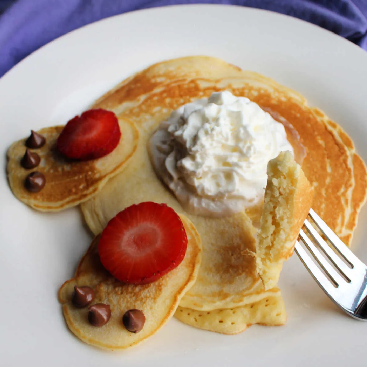 close up showing texture of homemade pancakes with bite on fork, whipped cream bunny tail and strawberry and chocolate chip bunny feet decorations visible in background.