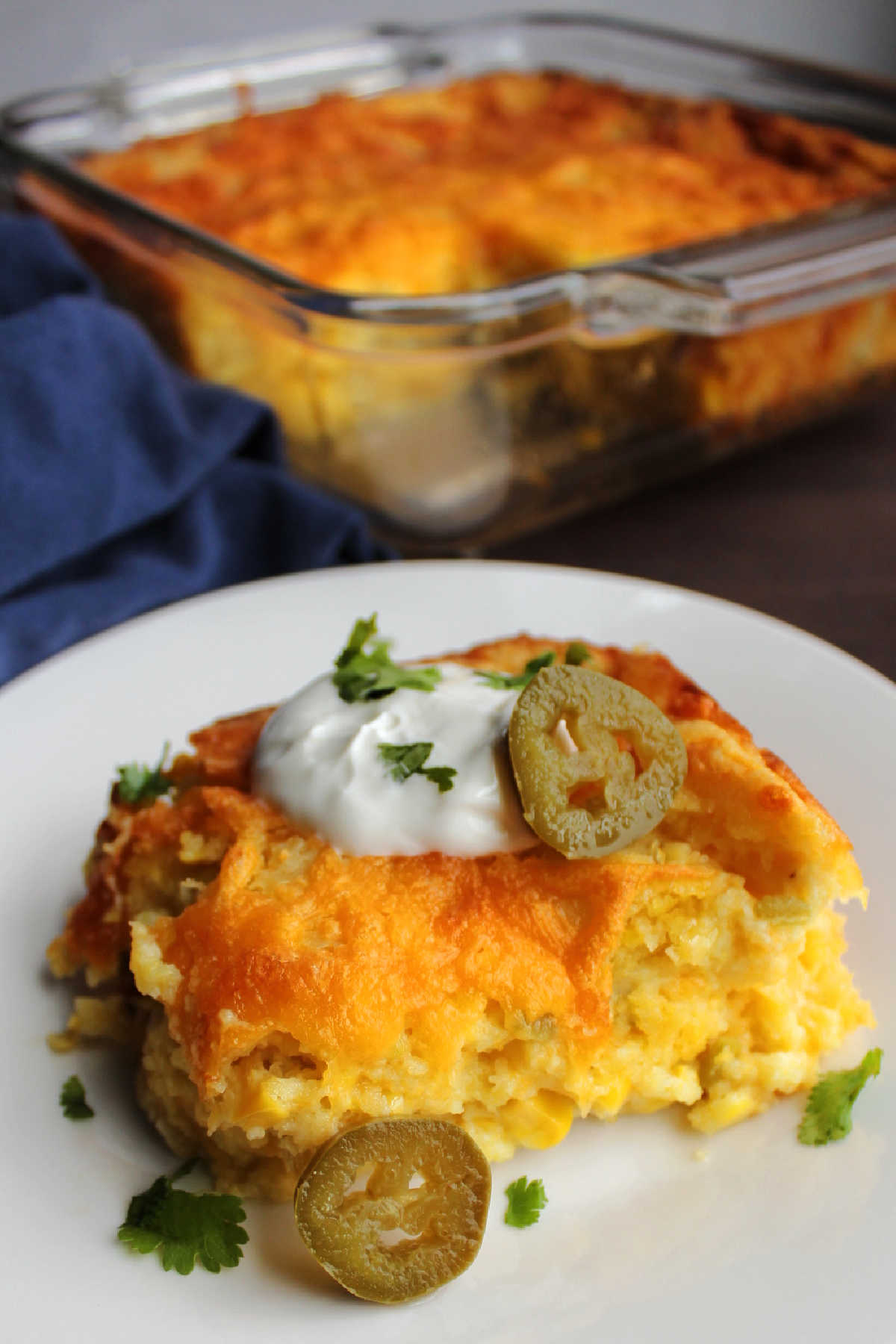 Serving of corn casserole with green chiles and cheddar cheese topped with sour cream and jalapenos in front of pan.