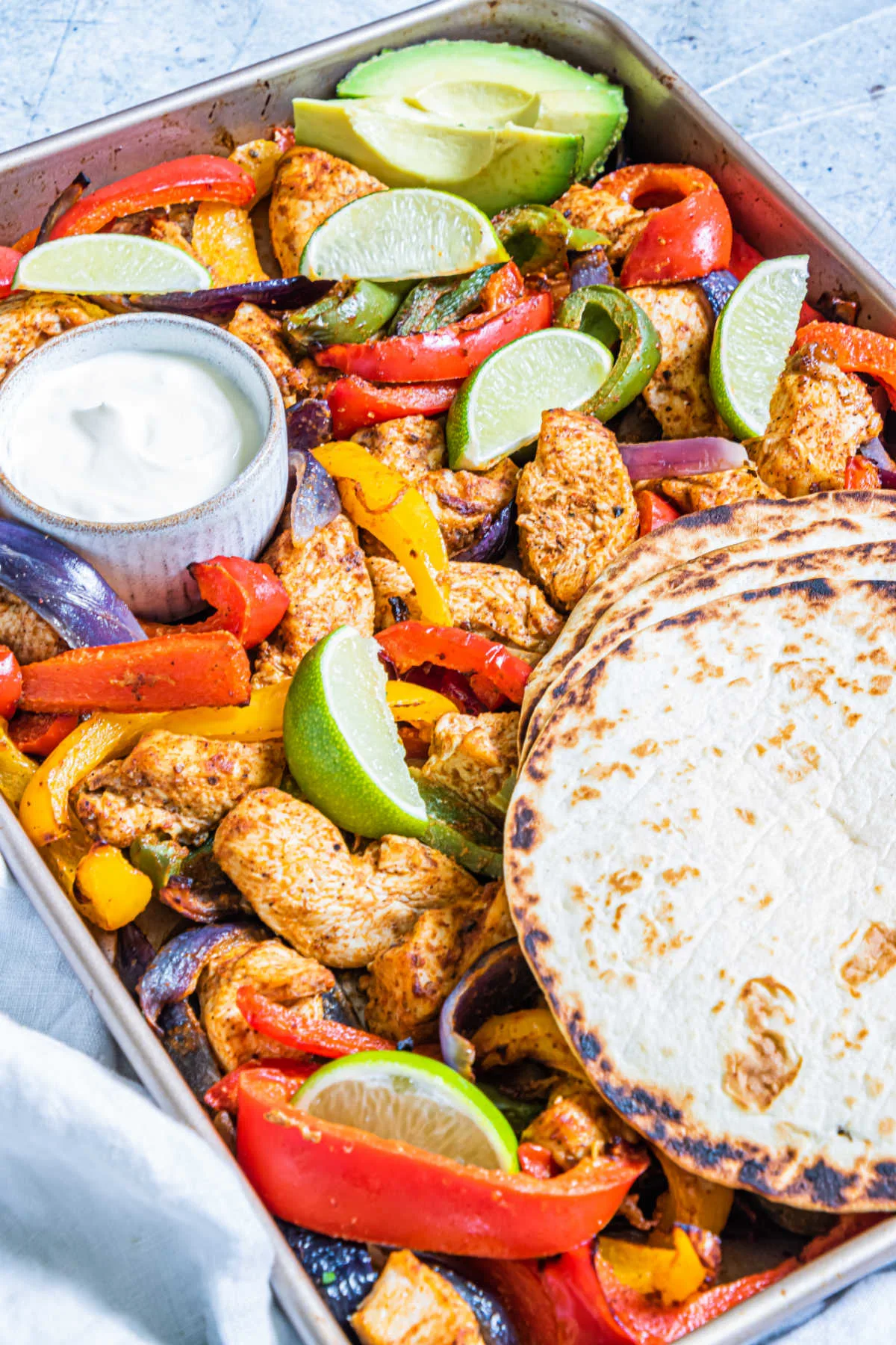 sheet pan filled with chicken fajitas with warm tortillas, lime sedges and a ramekin of sour cream.
