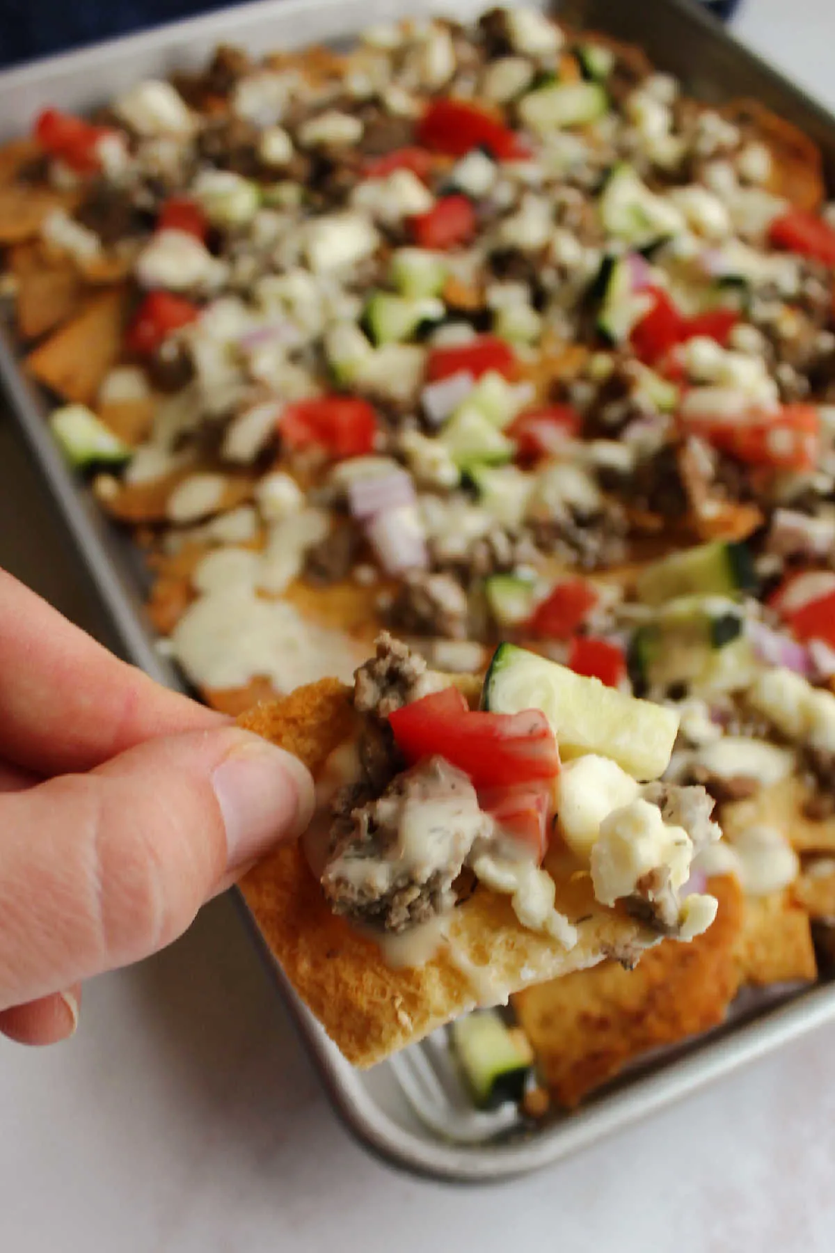 Hand holding pita chip topped with meat, cucumber, tomatoes, feta and tzatziki sauce.