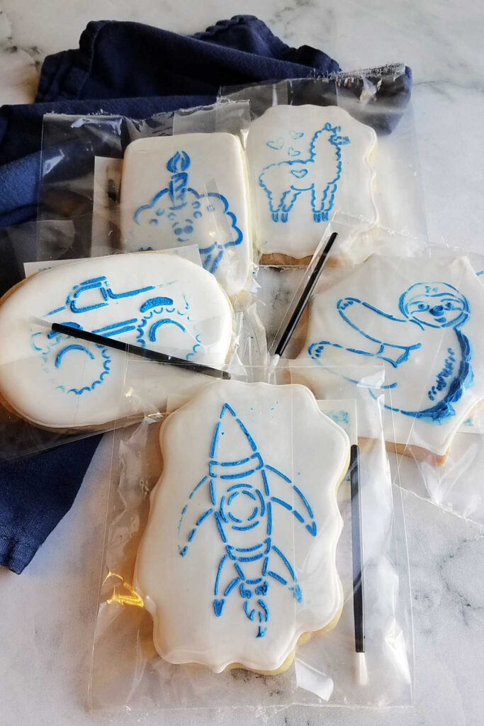 Packages of sugar cookies with white royal icing, blue royal icing outlines of shapes, small paintbrushes and edible watercolors to decorate them.