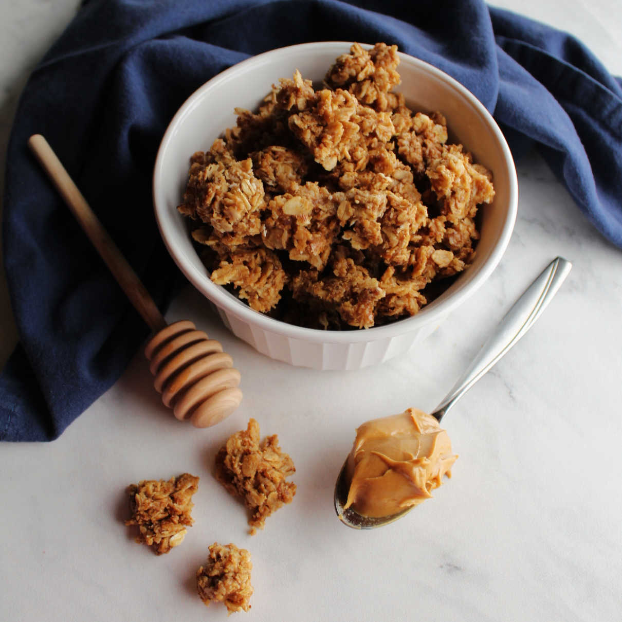 bowl of peanut butter granola next to a spoon of peanut butter and a honey drizzler.