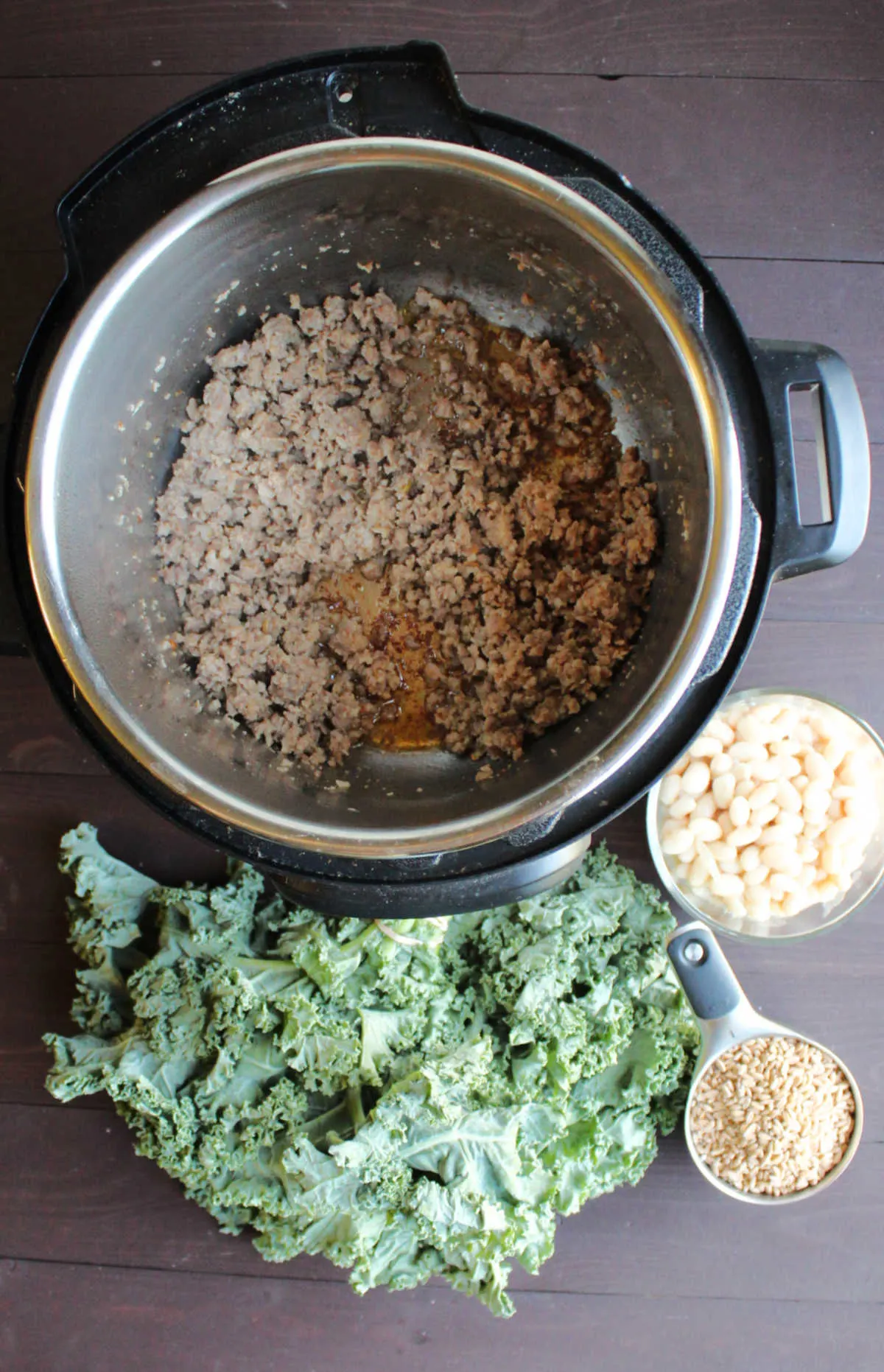 Browned Italian sausage in instant pot next to bunch of kale, bowl of white beans and cup of farro.
