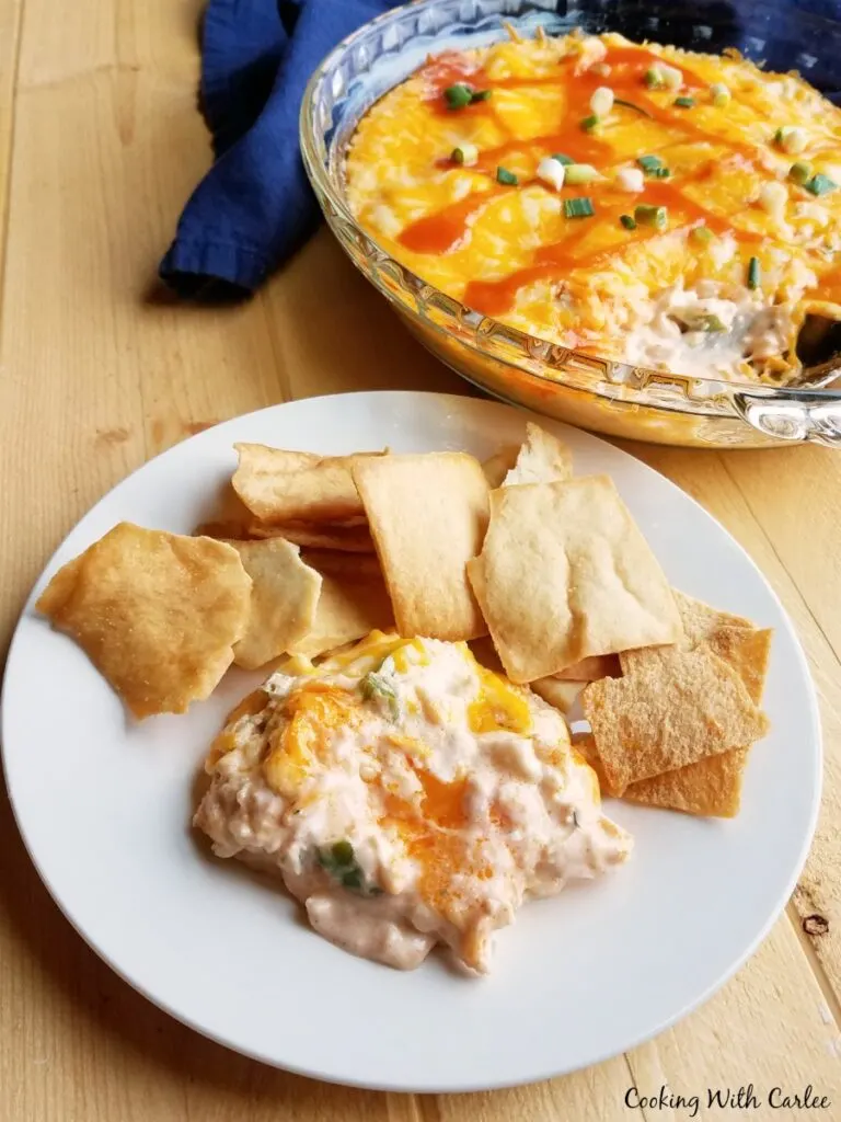 Serving of warm melty buffalo chicken dip on plate with pita chips in front of pan of remaining dip.