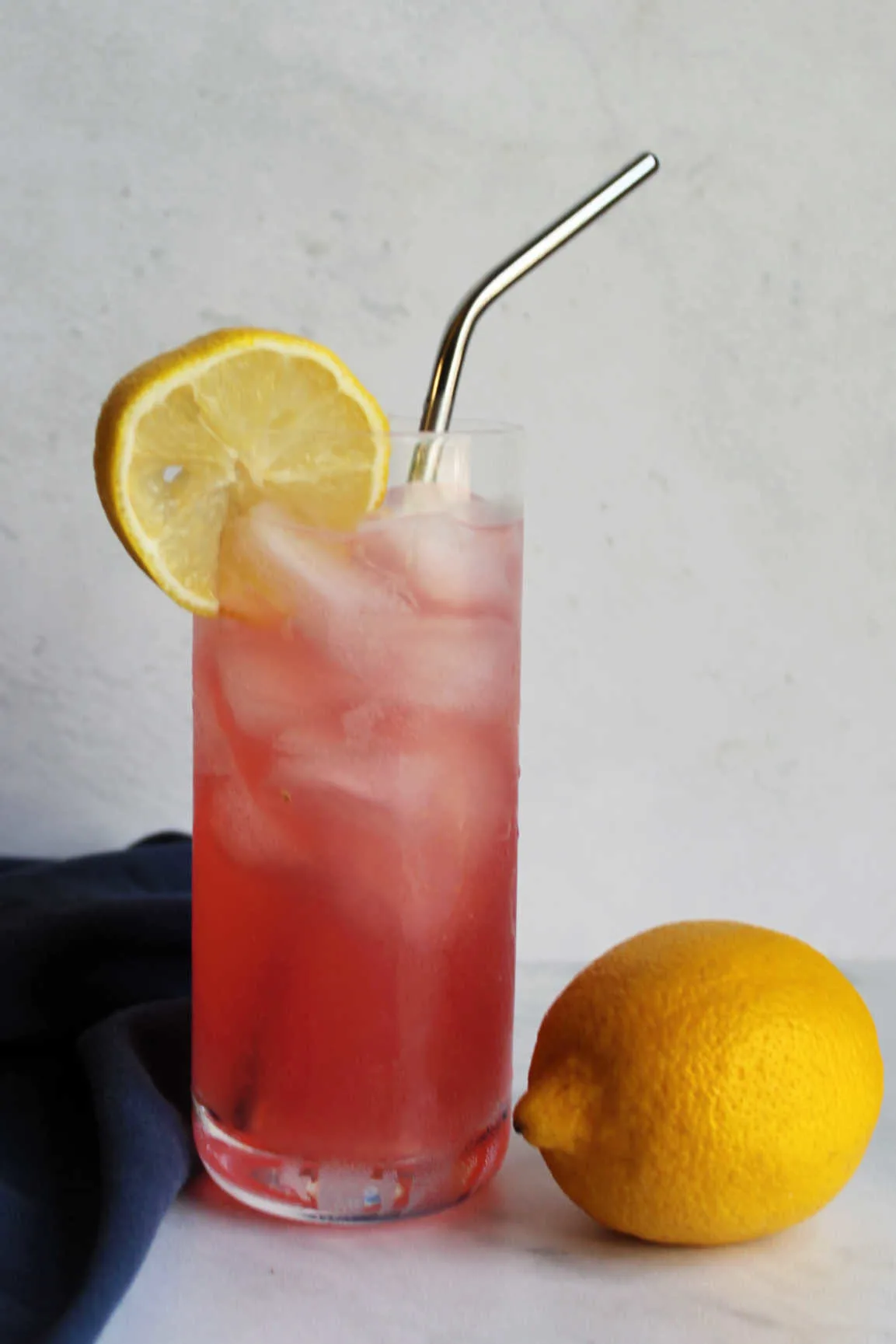 Single glass of pink lemonade in tall collins glass with metal straw next to a whole lemon.