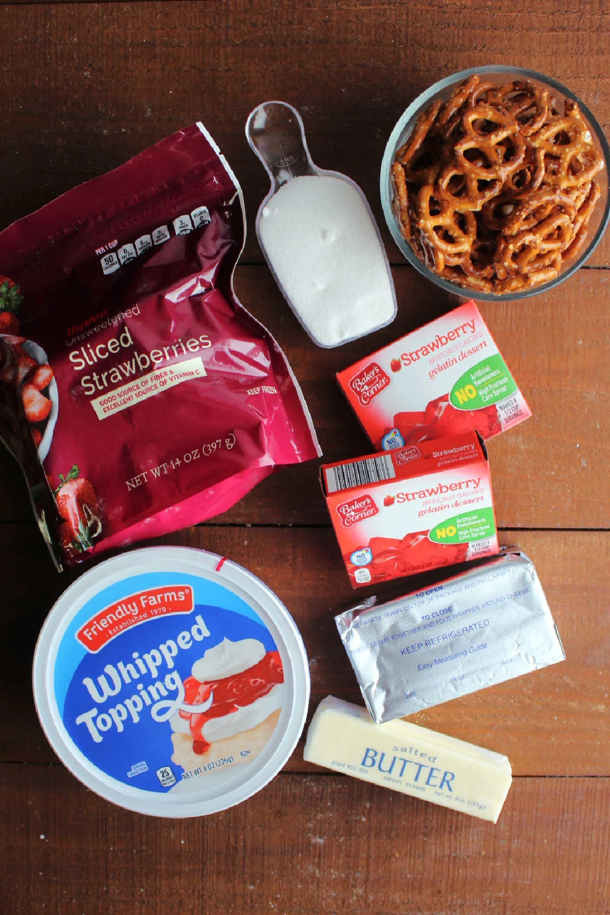 Ingredients including pretzels, sugar, butter, strawberry jello, frozen sliced strawberries, and whipped topping ready to be made into strawberry pretzel dessert.