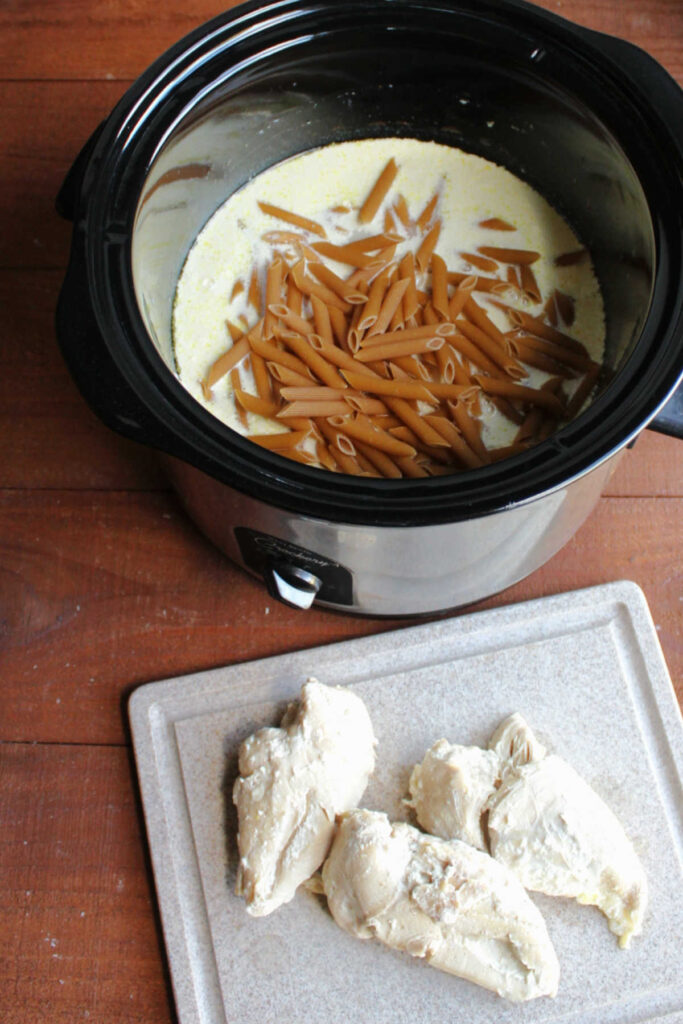Whole wheat penne in crock pot with creamy mixture and chicken nearby.