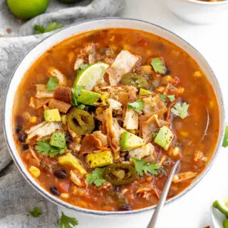 Bowl of chunky chicken tortilla soup topped with tortilla strips, jalapenos, avocado, a lime wedge and cilantro.
