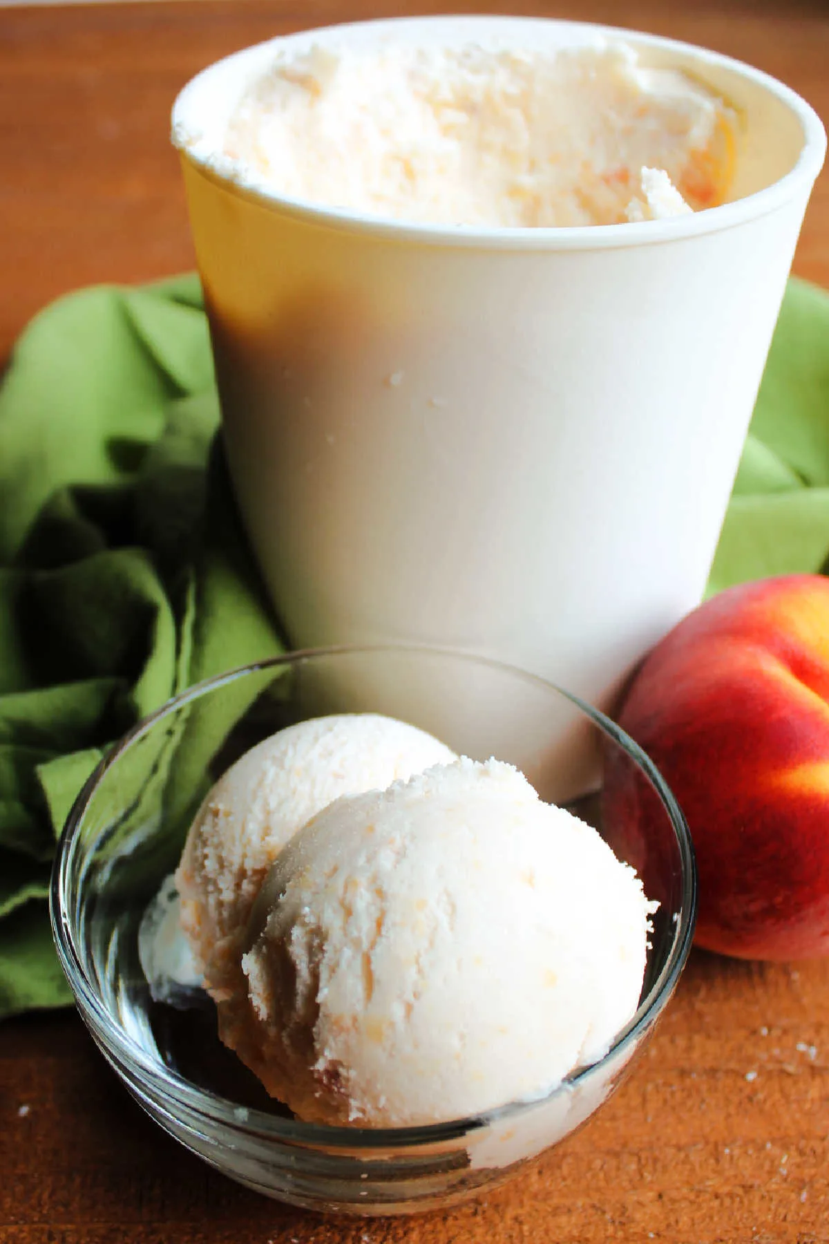 Small bowl of peach ice cream next to a fresh peach and an ice cream storage container.