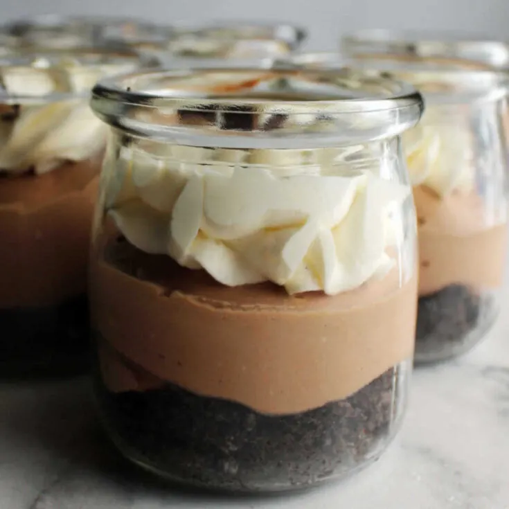 small glass jar with layer of chocolate crumb crust, creamy chocolate middle and piped ruffly white topping with mini chocolate chips on top.