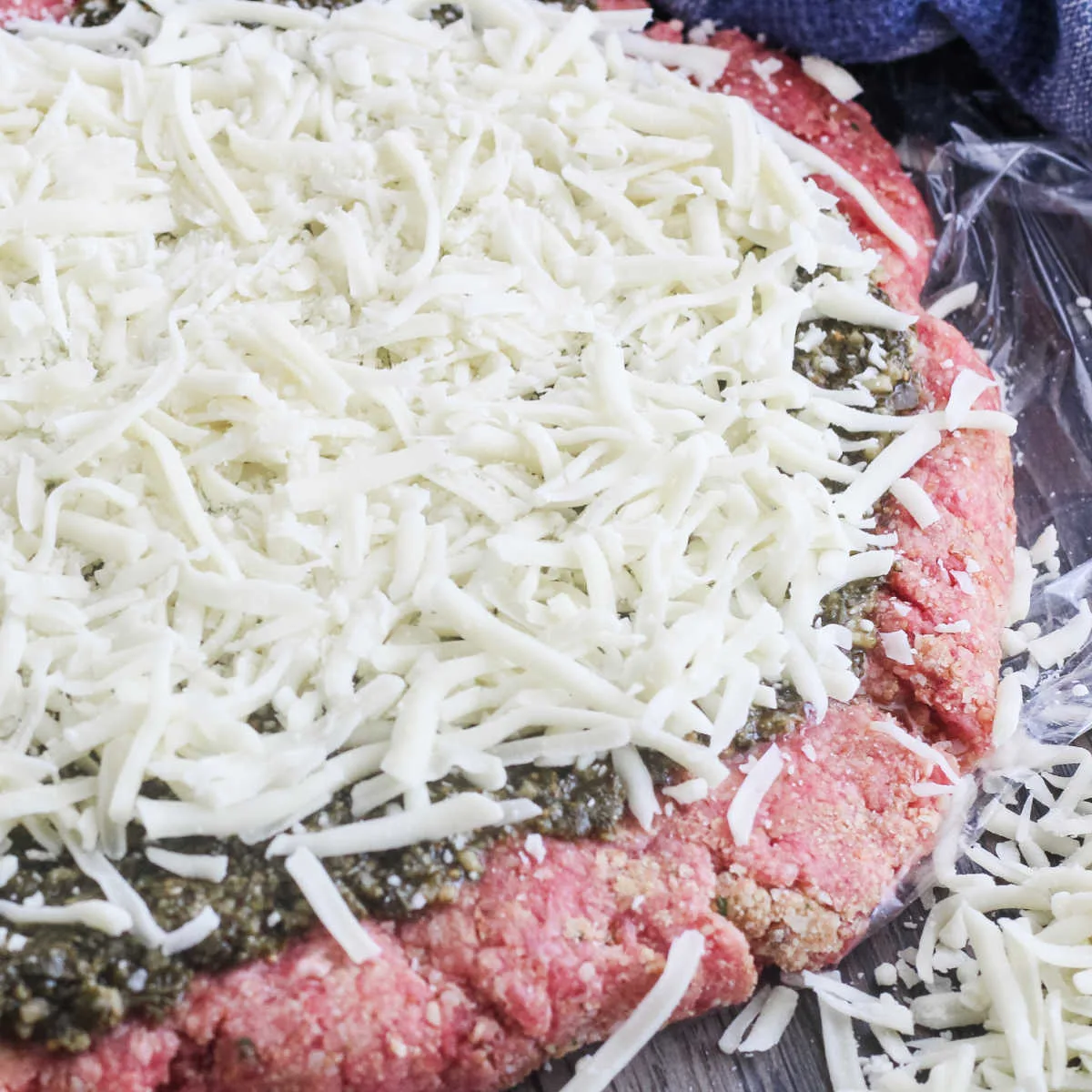 Layer of ground beef covered with pesto and cheese ready to be rolled into meatloaf.