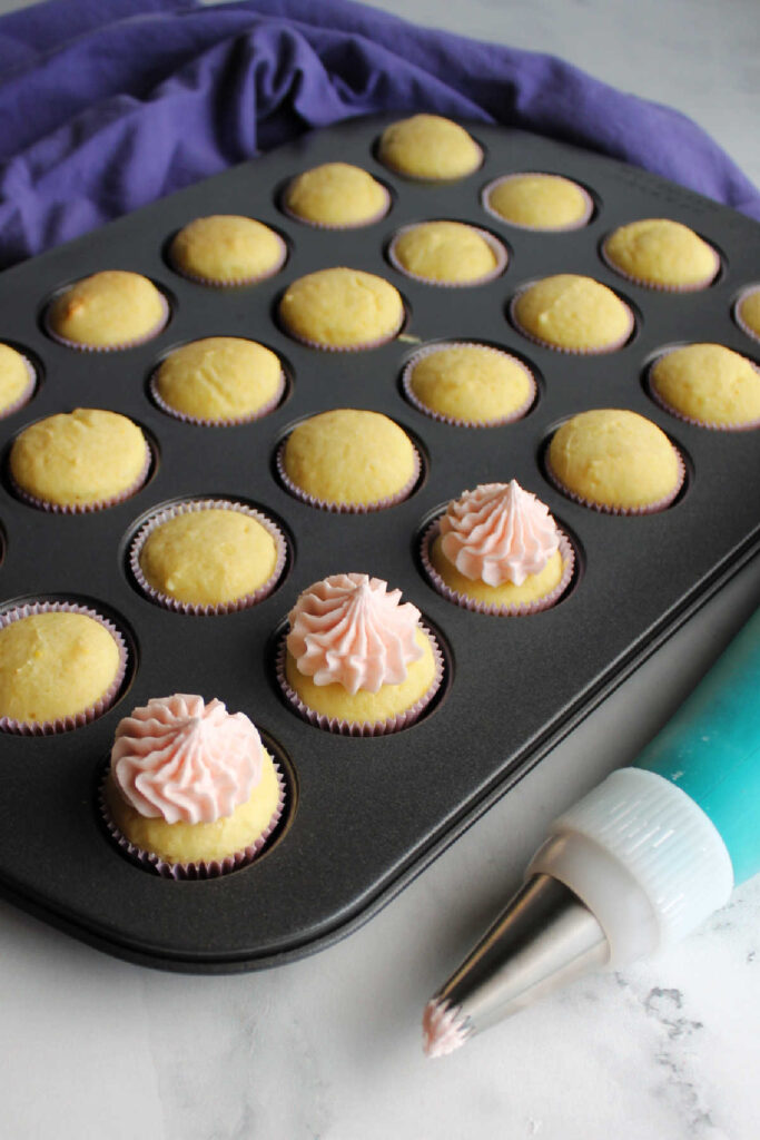 Piping bag next to pan of mini cupcakes some with pink lemonade frosting on top.