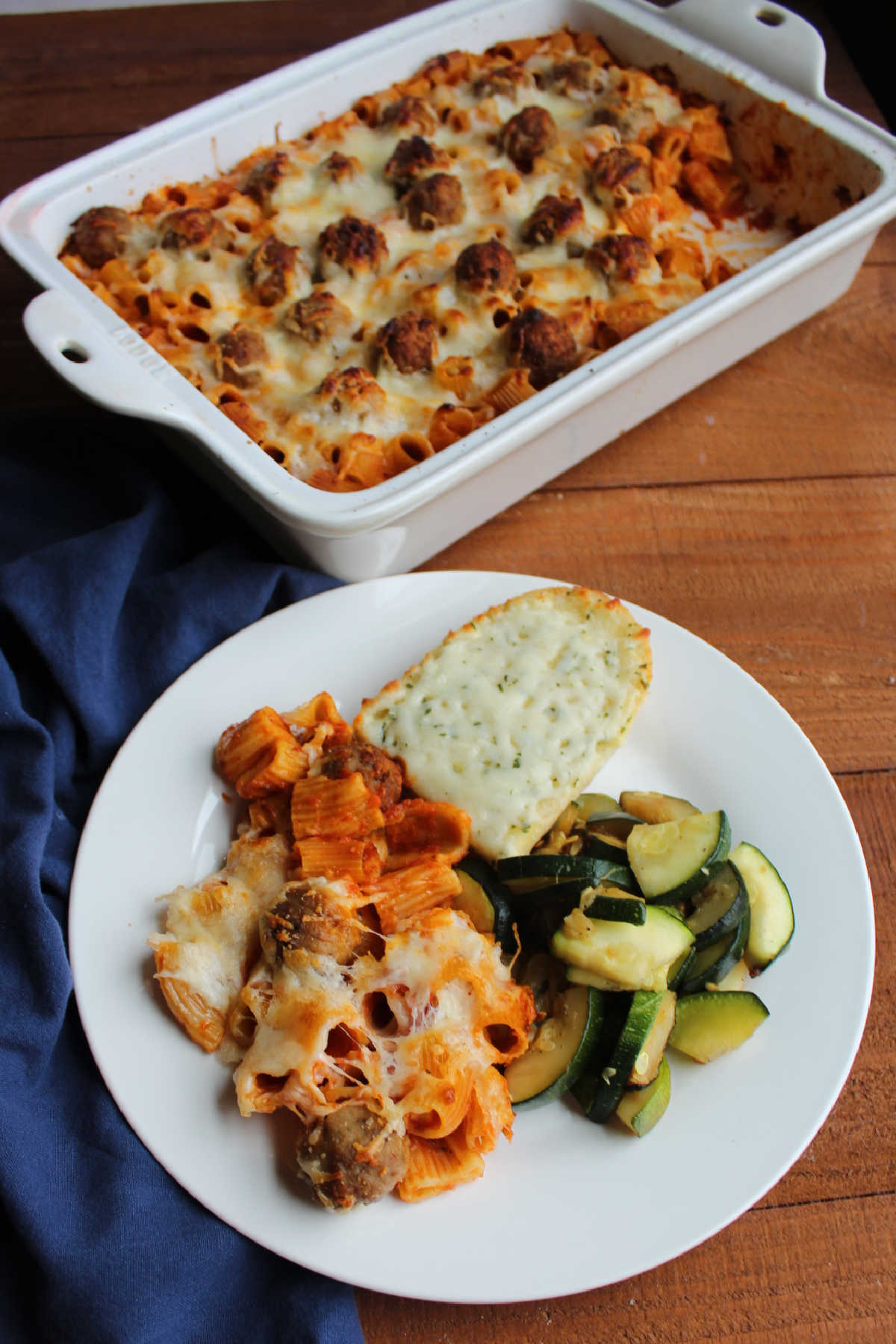 Plate of rigatoni and meatball casserole with garlic bread and zucchini next to remaining casserole.