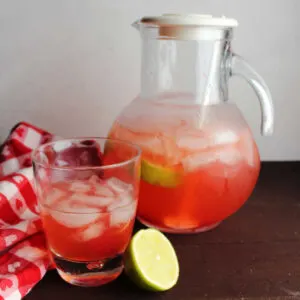 Pitcher of pink cherry limeade next to glass of limeade and a lime half.