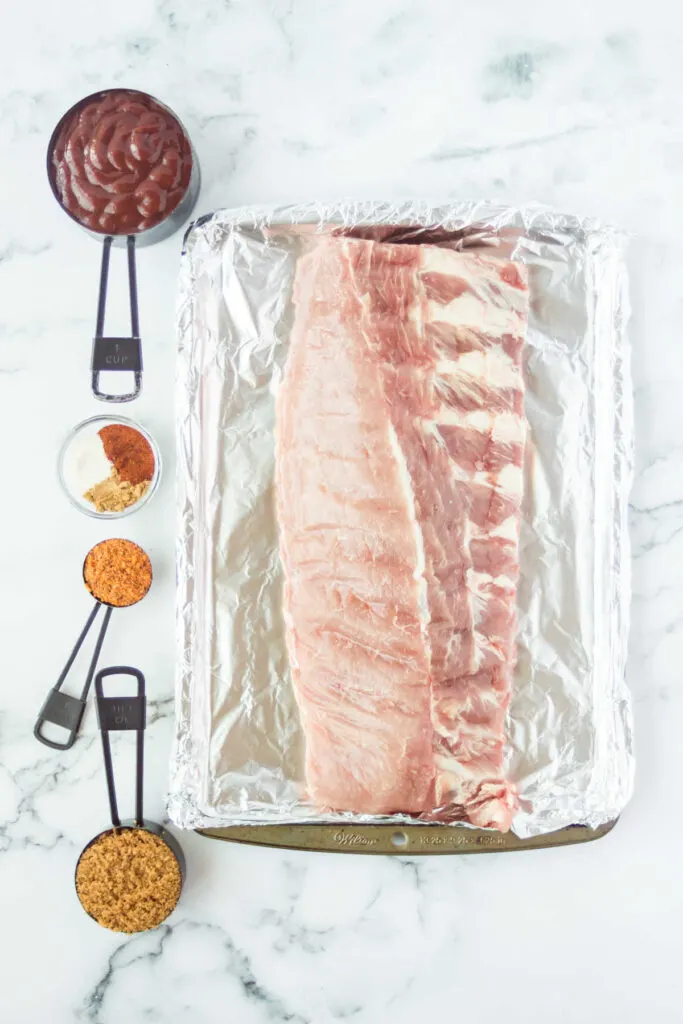 Rack of ribs on foil lined pan with seasonings and sauce nearby.