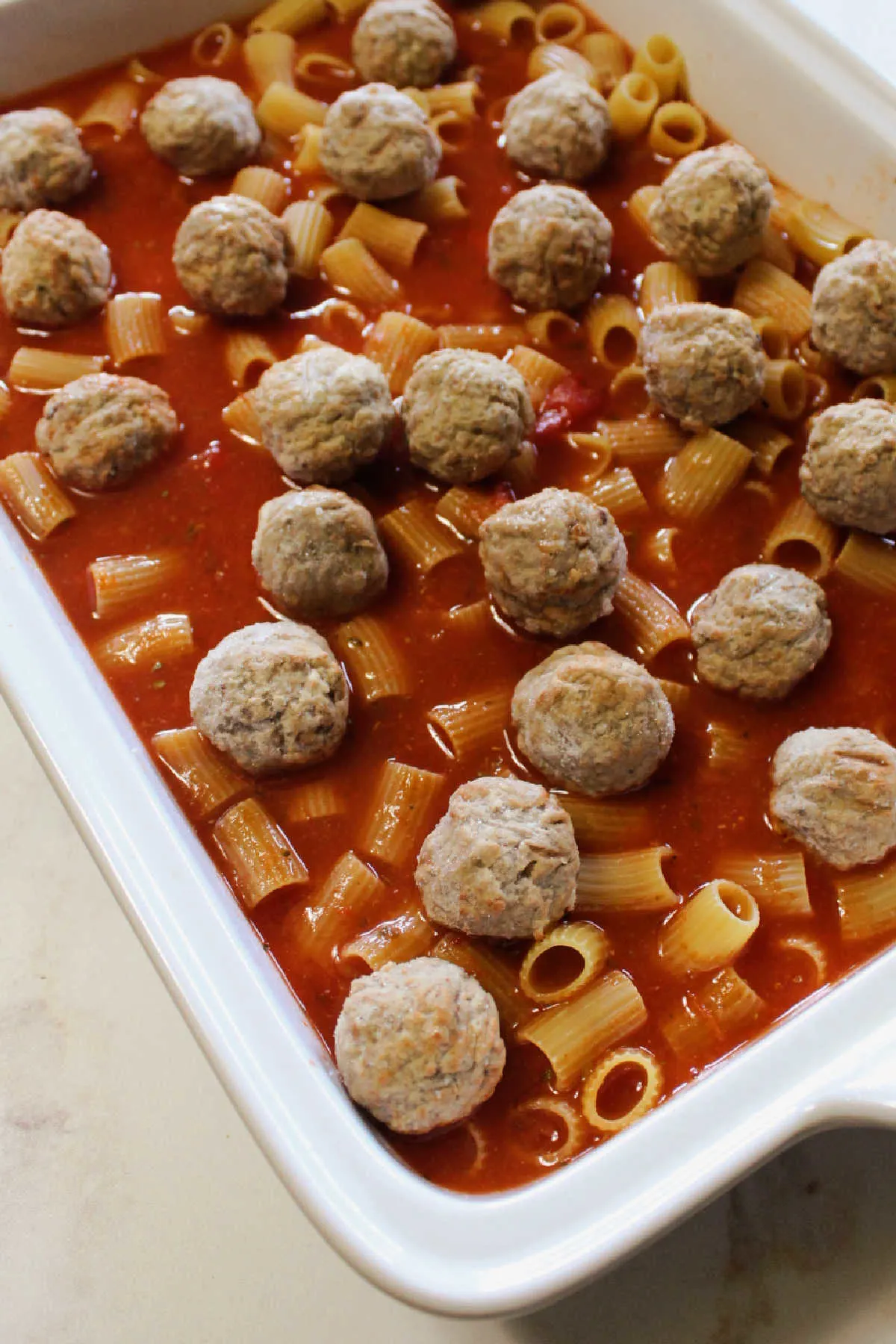 Uncooked pasta and tomato sauce in dish with frozen meatballs ready for the oven.