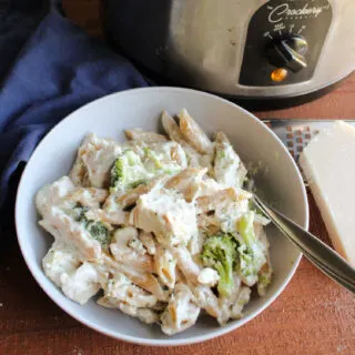 Bowl of penne pasta with chicken, broccoli and creamy alfredo sauce next to crock pot.