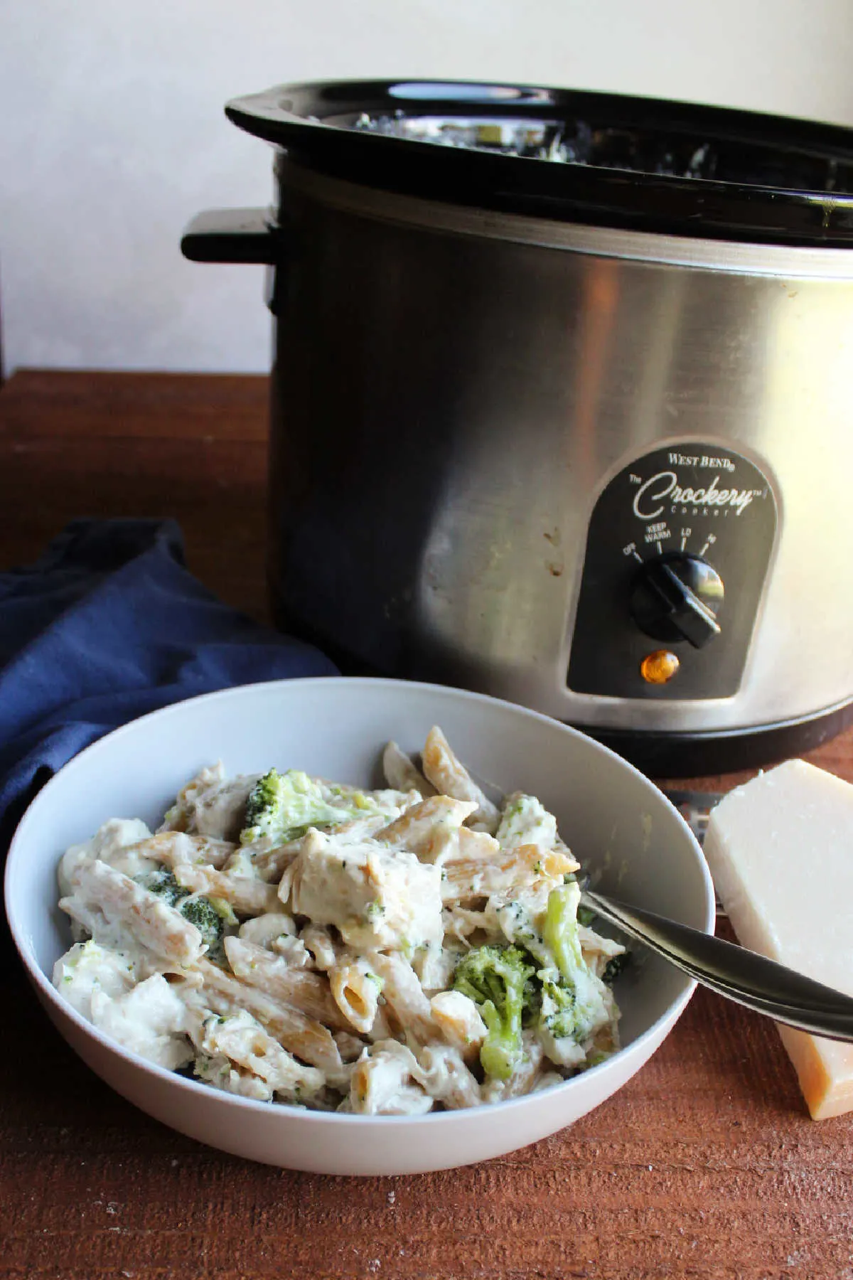 Bowl of pasta, chicken and broccoli coated in alfredo sauce in front of a crock pot.