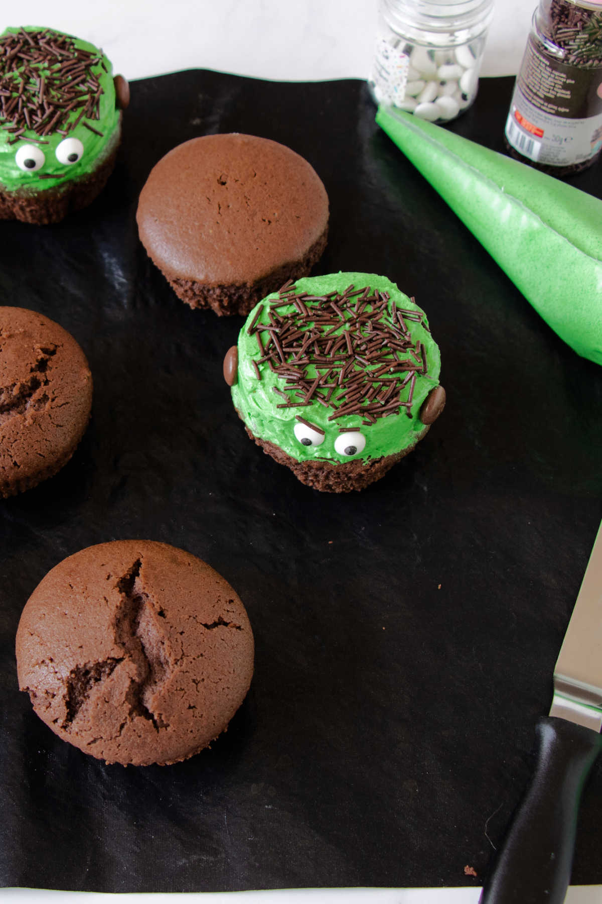 Freshly baked chocolate cupcakes with piping bag filled with green buttercream, small thing candy eyes and chocolate sprinkles.