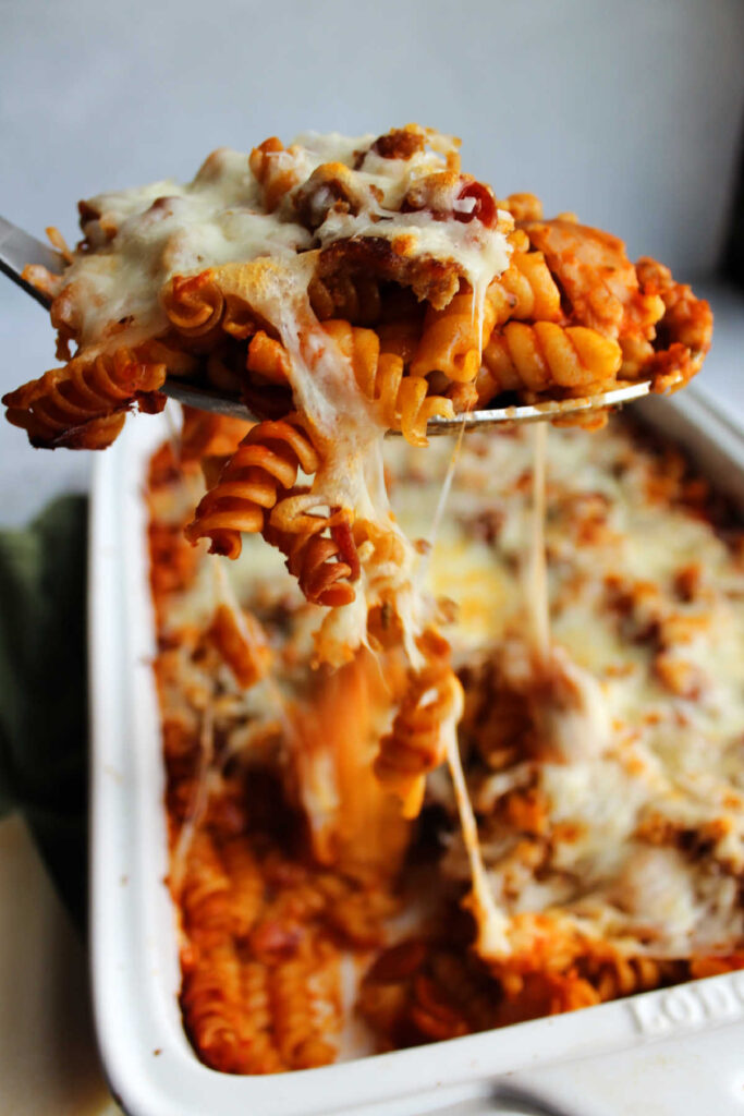 Serving spoon lifting out helping of pizza pasta bake with strings of melted mozzarella cheese pulling from dish.