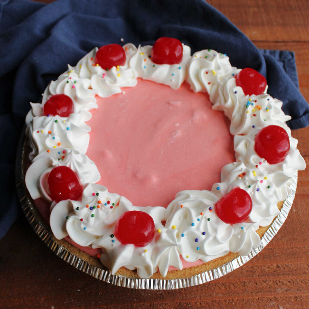 Whole pink cherry jello pie with whipped topping piped around the edge and maraschino cherries on top.