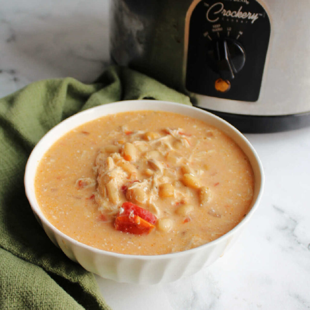 Bowl of creamy soup with white beans, chicken and bits of tomato showing in front of crock pot. 