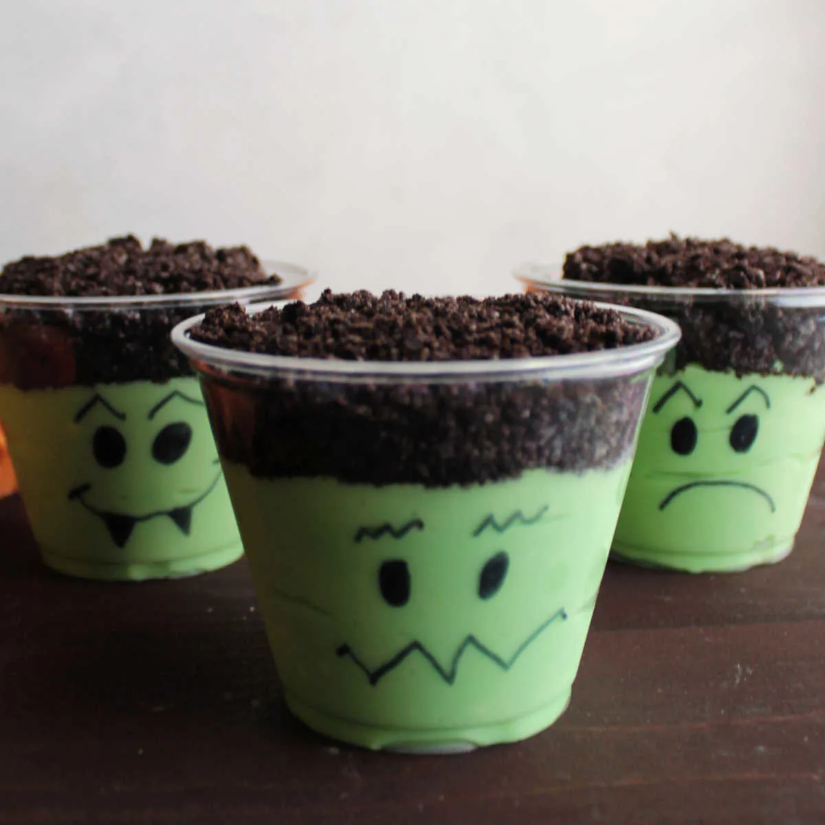 Three pudding cups with green pudding mixture and chocolate cookie crumb topping with faces drawn on them to look like Frankenstein.