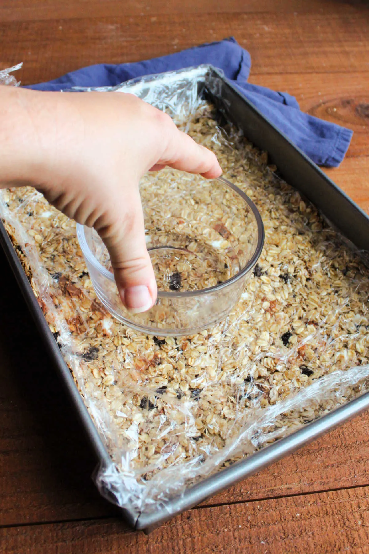 Pressing down on homemade granola bars with bowl on top of plastic wrap.