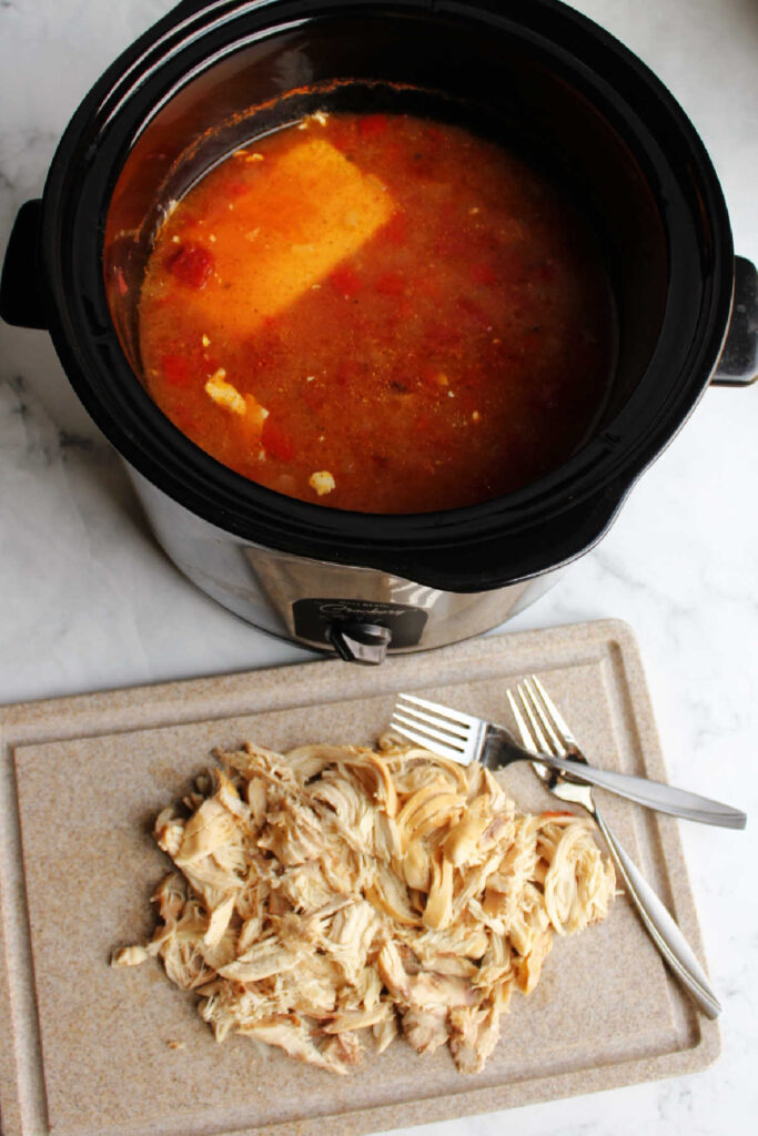 shredded chicken on cutting board in front of slow cooker with soupy broth.