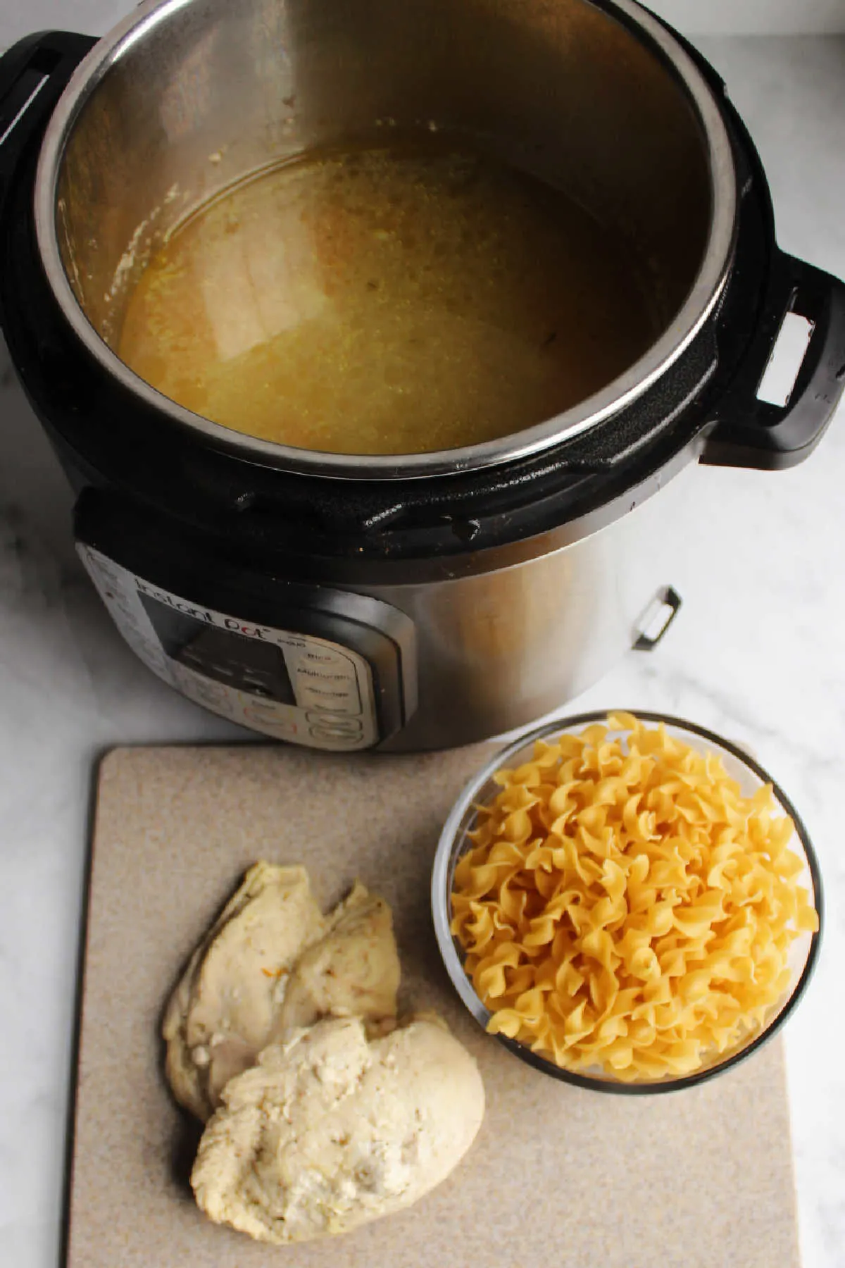 Cooked chicken on cutting board with bowl of raw pasta next to instant pot of broth waiting for pasta.