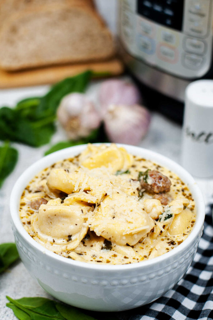 Bowl of creamy soup with cheese tortellini, chunks of Italian sausage and spinach inside sitting in front of instant pot.