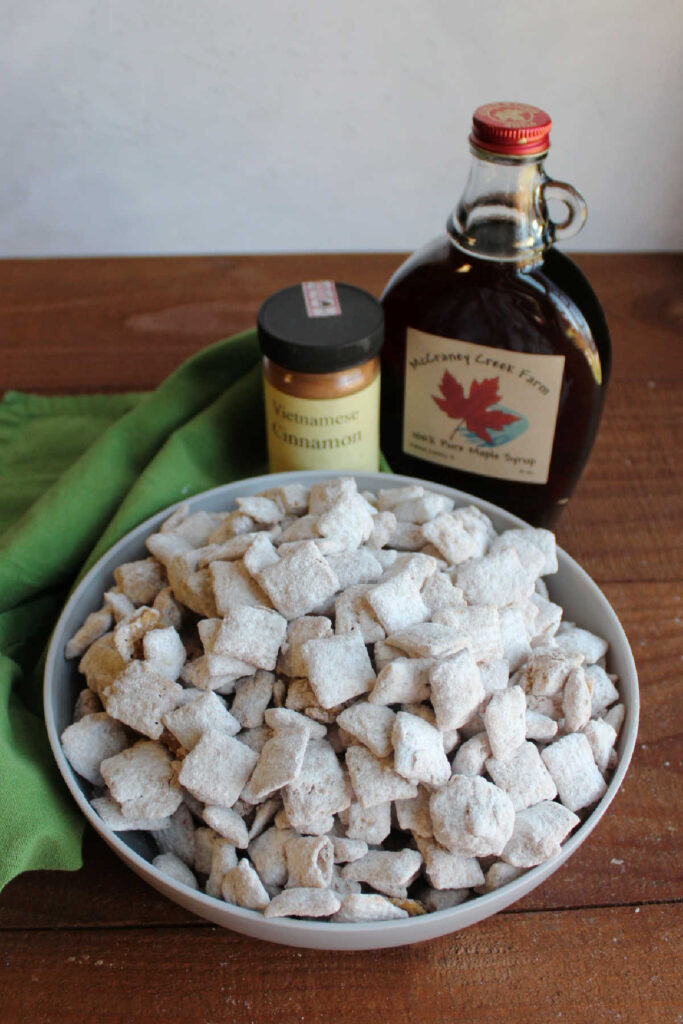 Bowl of maple cinnamon puppy chow next to bottle of pure maple syrup and jar of cinnamon.