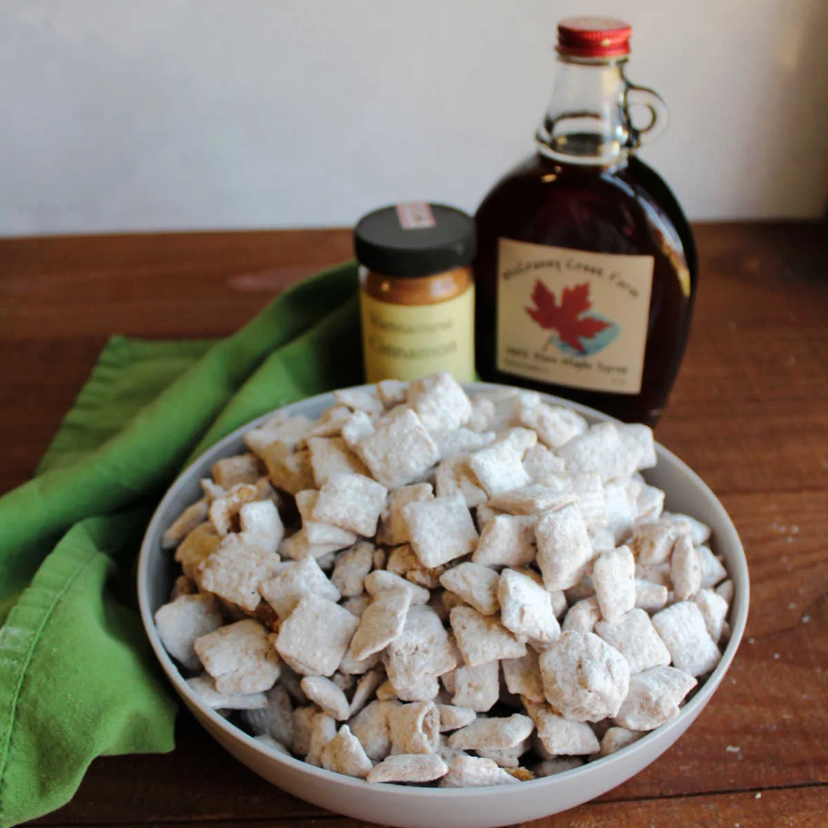 Bowl of powdered sugar coated french toast puppy chow in front of bottle of maple syrup and cinnamon.