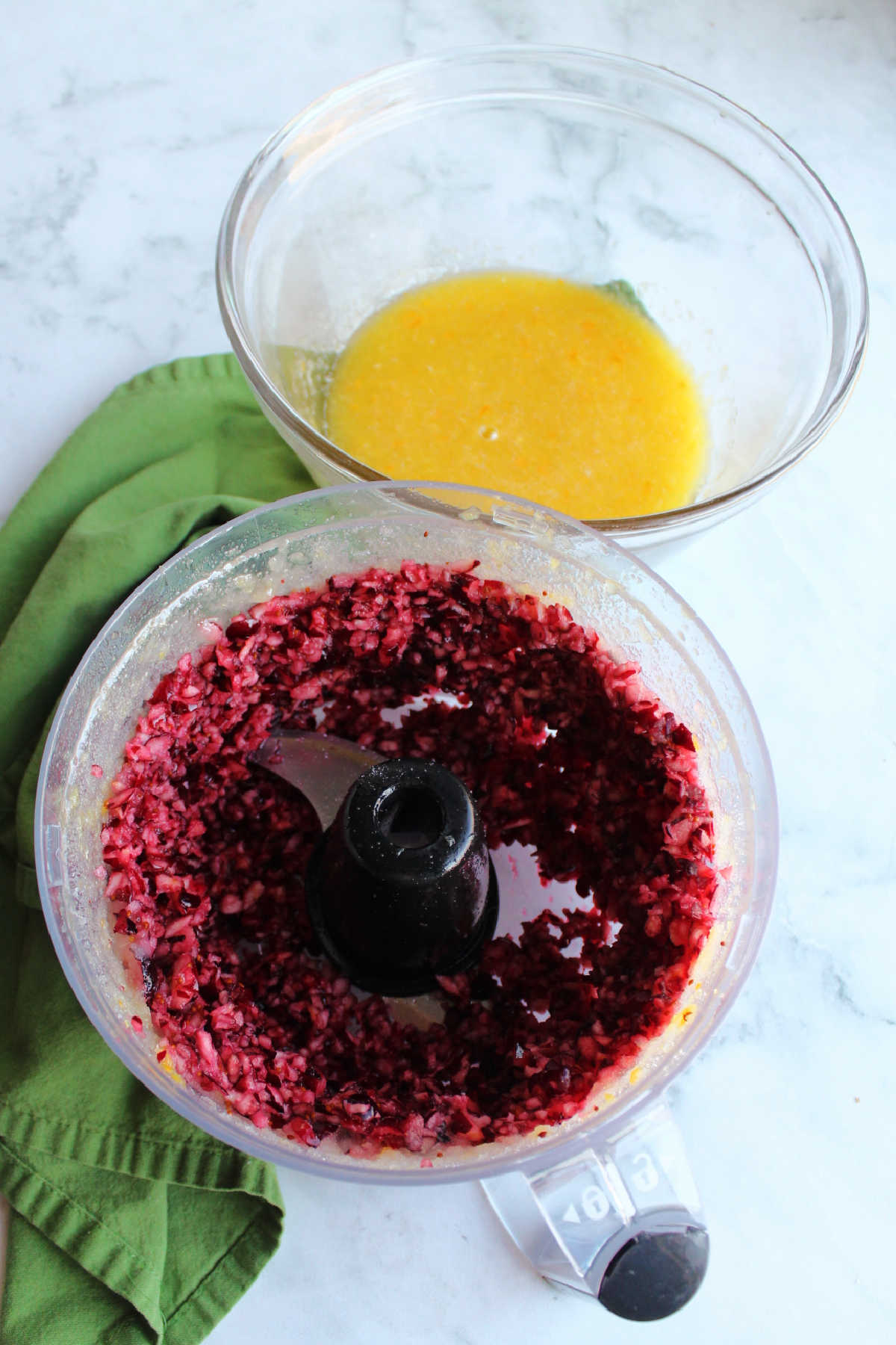 Food processor filled with bits of cranberry next to bowl of orange and sugar mixture.