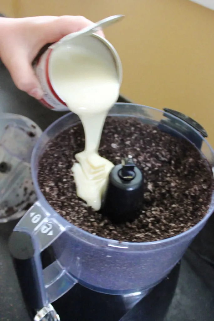 Pouring can of condensed milk into food processor filled with Oreo crumbs.