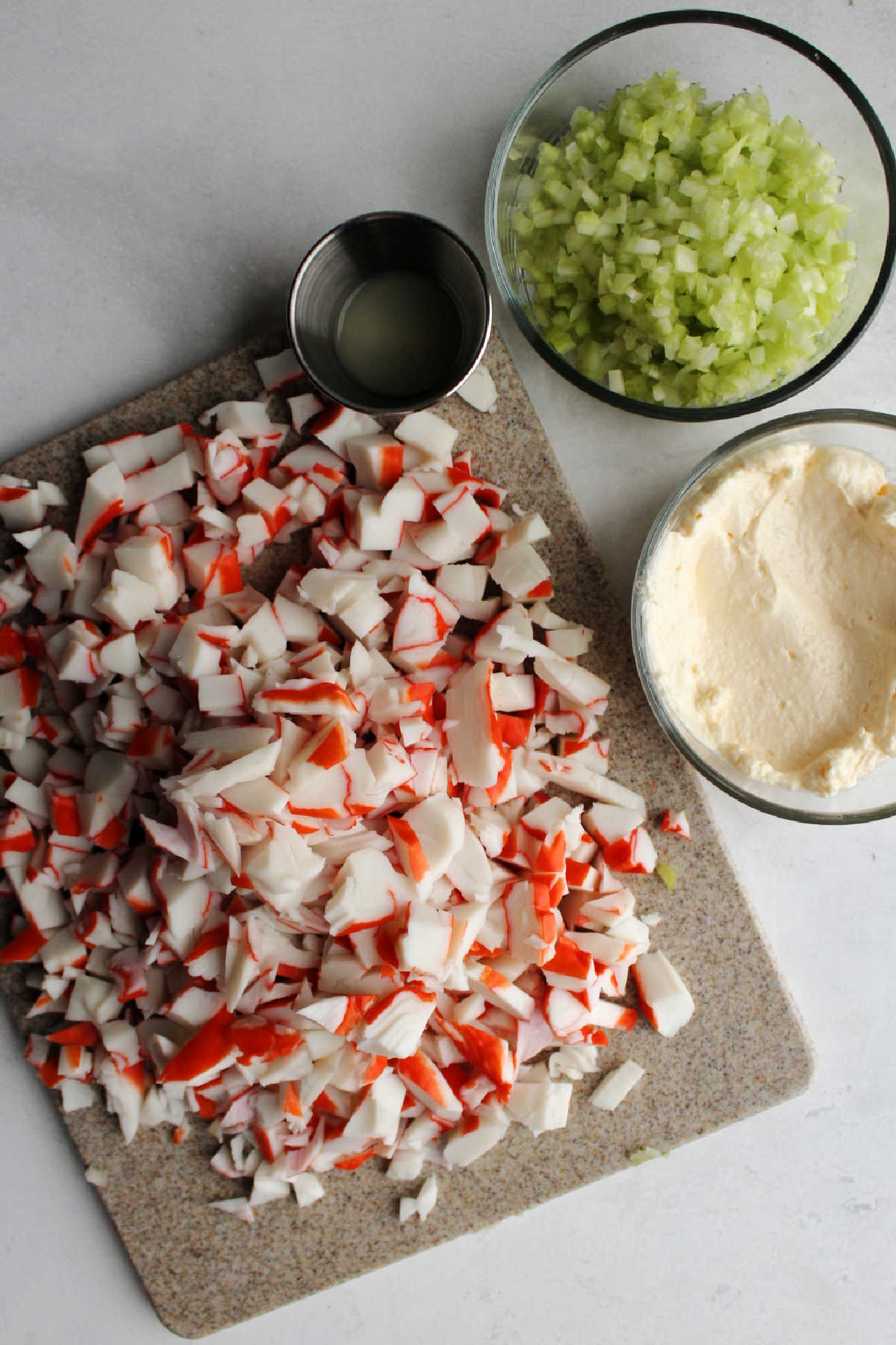 Cutting board filled with chopped crab next to bowls of mayonnaise, chopped celery and lemon juice.