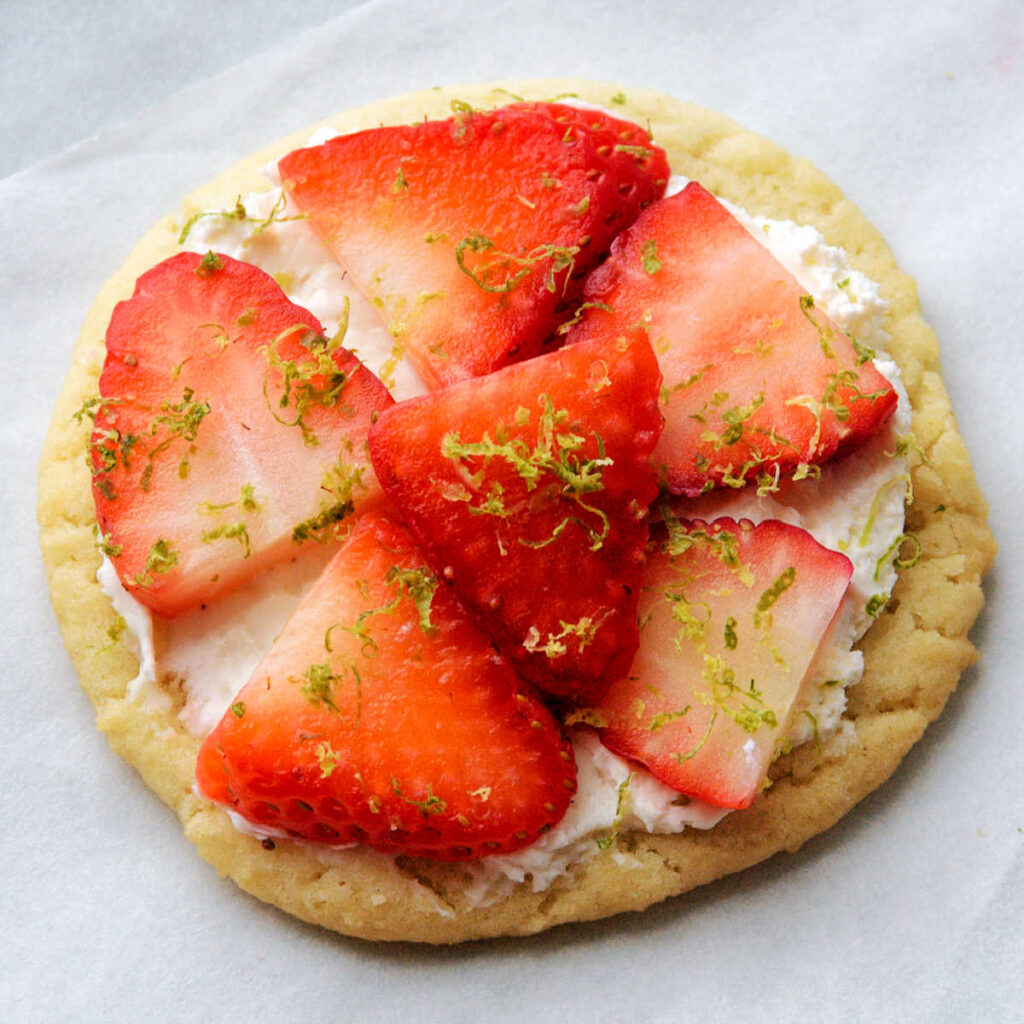 Small fruit pizza made from sugar cookie, cream cheese spread, sliced strawberries and lime zest. 