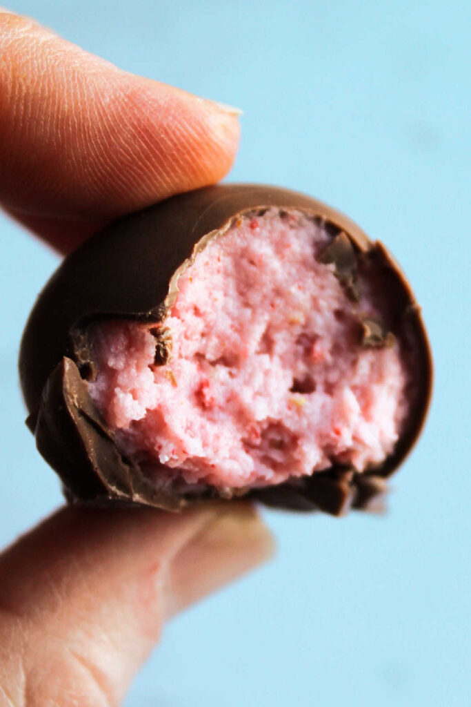 Hand holding truffle with a bite taking out, showing strawberry center surrounded by milk chocolate shell.