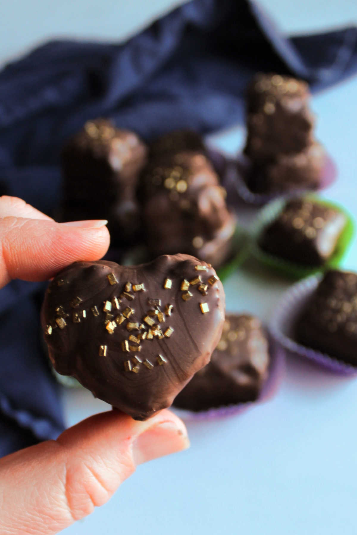 Hand holding heart shaped petit four dipped in dark chocolate and topped with gold sugar sprinkles.