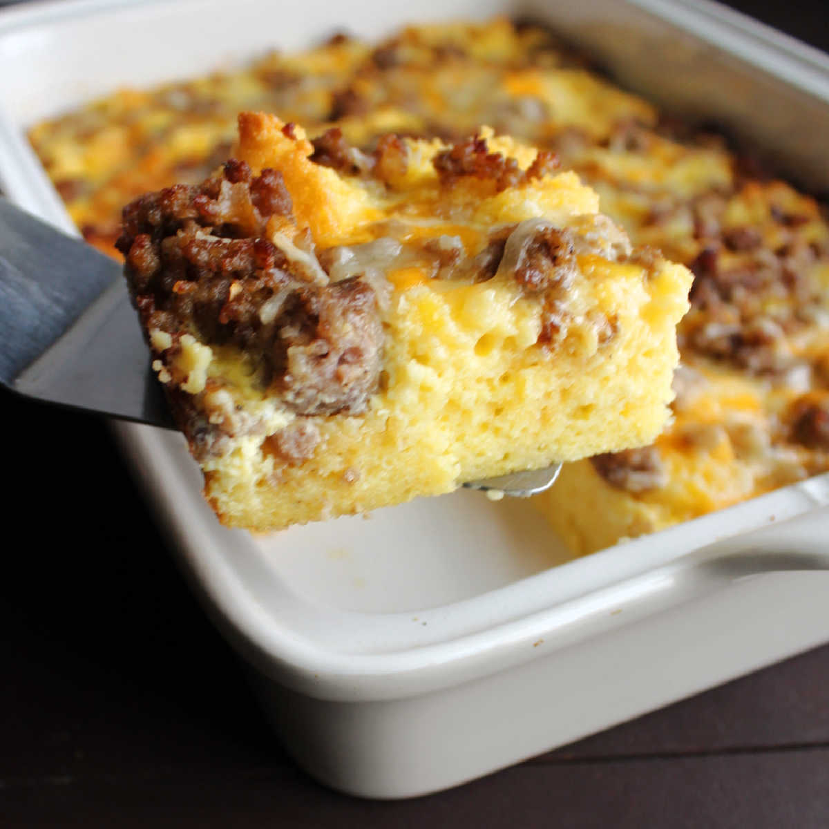 Spatula lifting out piece of egg casserole with cornbread, sausage and cheddar cheese.