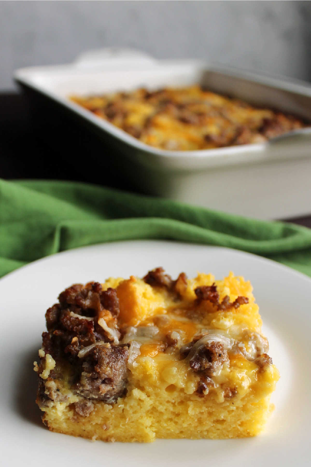 Piece of sausage, cheese, egg and cornbread casserole served in front of baking dish.