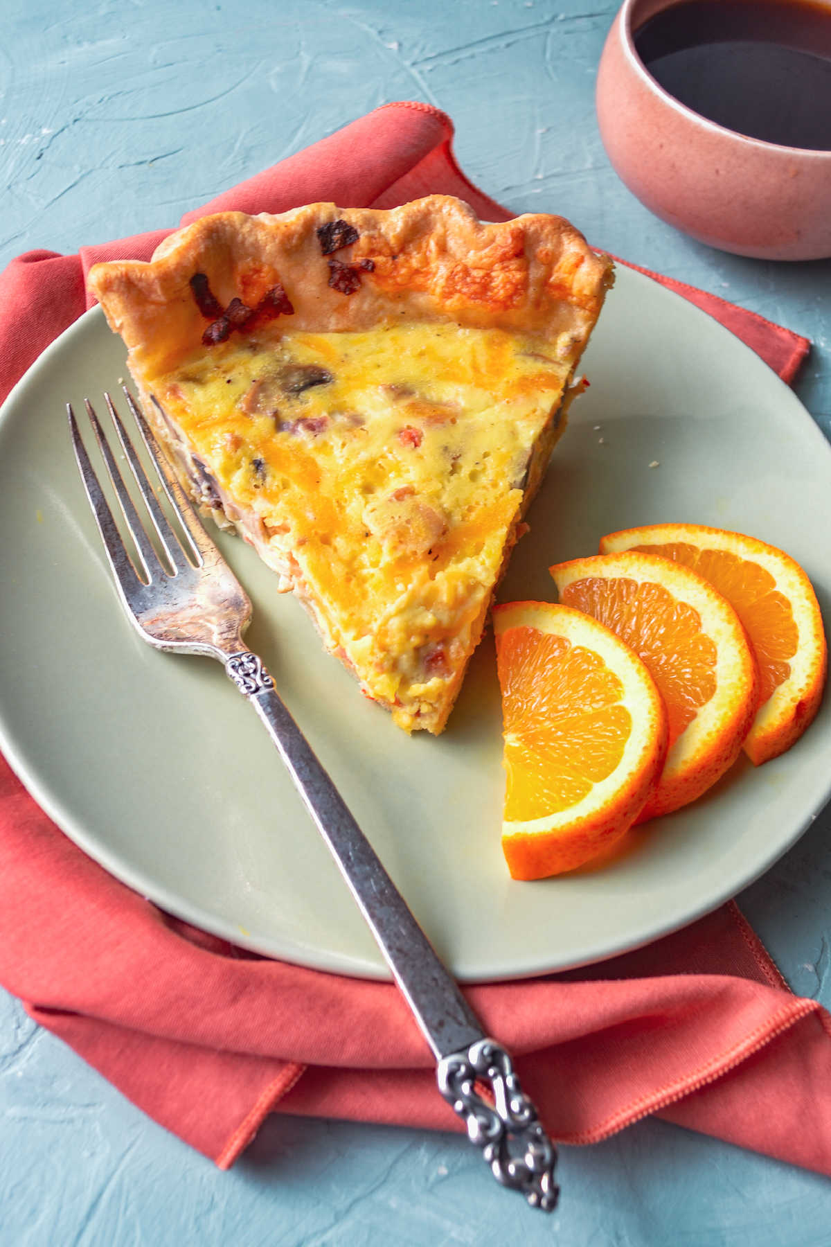 Breakfast plate with slice of quiche, fork orange slices and a cup of coffee ready to eat. 