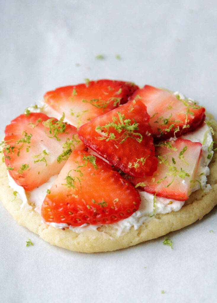 Close up of chewy sugar cookie topped with cream cheese and slices of fresh strawberry topped with lime zest.